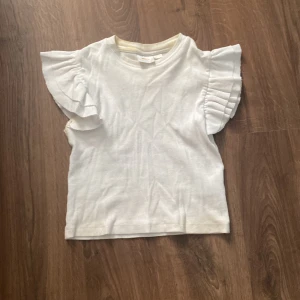 Vit topp med volangärmar från Gina Tricot - Säljer en söt vit topp från Gina Tricot i storlek 134/140. Den har fina volangärmar som ger en extra touch till outfiten. Perfekt för sommarens varma dagar eller som en söt detalj under en kofta. Toppen är i bra skick och redo för en ny ägare! Förutom att den har lite smink fläckar på insidan så syns det inget . Fläckarna är på insidan 