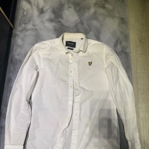 Vit skjorta från Lyle & Scott - Säljer en klassisk vit skjorta från Lyle & Scott i bomull. Den har långa ärmar och knappar framtill. På bröstet finns deras ikoniska logga i gult och svart. Perfekt för både vardag och fest! 🕺