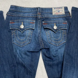 Blå jeans från True Religion - Säljer ett par snygga blå jeans från True Religion i bra skick! W36, inner ben längd 77 och lår bredd ca 24. Obs materialet är lite stretchit så även om lår bredden skulle vara lite för litet skulle du förfarande kunna använda dom. De har en tight passform och är lågmidjade med coola detaljer som knappar och broderade fickor. Perfekta för både vardag och fest!