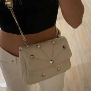 Beige quiltad axelväska med kedja - Snygg beige axelväska med quiltat mönster och coola nitar. Väskan har en elegant kedja som axelrem och är perfekt för både vardag och fest. Den är i mycket bra skick och ger en lyxig touch till vilken outfit som helst.