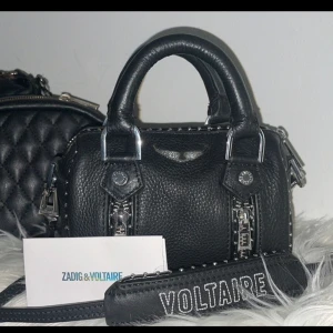 Svart handväska från Zadig & Voltaire - Snygg svart handväska i skinn från Zadig & Voltaire. Väskan har coola dragkedjedetaljer och en justerbar axelrem med texten 'VOLTAIRE'. Perfekt för både vardag och fest! säljer den på grund av att jag bara haft den som prydnad nästan aldrig använd känner att det kanske är någon annan som har bättre användning för den än än jag❤️ 