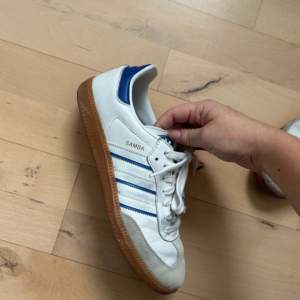 Säljer ett par klassiska Adidas Samba sneakers i vitt och blått. Skorna har en snygg retrodesign med bruna sulor och är i bra skick. Perfekta för både vardag och träning. De har snörning och är gjorda i skinn och syntetmaterial.