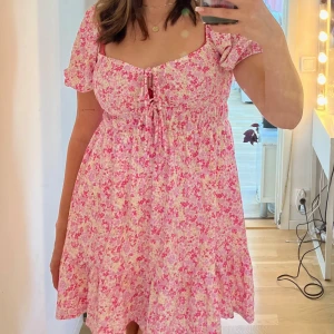 Blommig sommarklänning  - Söt blommig klänning i rosa och vit med korta ärmar och volangdetaljer. Perfekt för sommarens fester eller en dag på stan. Klänningen har en knytning framtill och en luftig passform som gör den bekväm att bära hela dagen. Använd 1 gång på midsommar