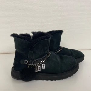 Svarta UGG boots med kedjedetalj - Säljer ett par svarta UGG boots i mocka med fluffigt foder. De har en cool kedjedetalj med UGG-logga och en söt boll på sidan. Perfekta för att hålla fötterna varma och stiliga under vintern. Använda men i bra skick.