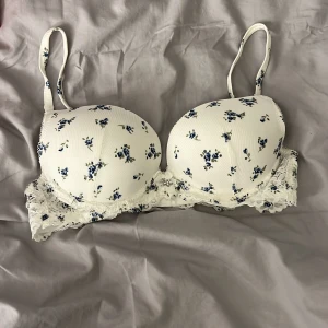 Vit blommig bh med spetsdetaljer - Söt vit bh med små blå blommor och spetsdetaljer i storlek 70D, kommer från hunkemöller och är aldrig använd