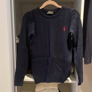 Mörkblå stickad tröja från Ralph Lauren - Säljer en snygg mörkblå stickad tröja från Ralph Lauren med det klassiska röda logotypbroderiet på bröstet. Tröjan har ett kabelstickat mönster och långa ärmar, perfekt för kyligare dagar. Passar både till jeans och chinos för en stilren look.