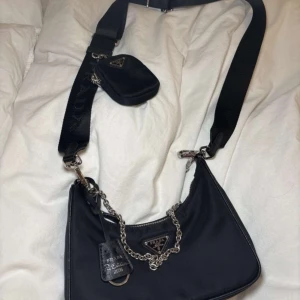 Svart axelväska från Prada - Snygg svart axelväska från Prada med silverkedja och matchande liten väska. Väskan har en klassisk design med dragkedja och det ikoniska Prada-emblemet på framsidan. Perfekt för att ge en stilren touch till din outfit.