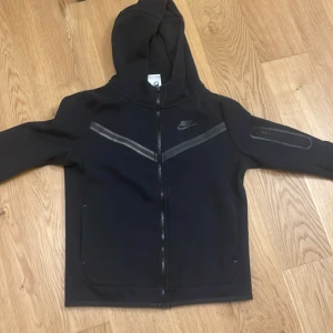 En snygg svart Nike Tece fleece - Säljer en svart huvtröja från Nike med dragkedja och huva. Tröjan har långa ärmar och en diskret logga på bröstet. Perfekt för en sportig/avslappnad look. Nypris 1099kr. Skick 7-10 pågrund av att hela hoodie är normal men som man ser på bild 1 har ett av strecken förlorat lite färg. Pris kan diskuteras!
