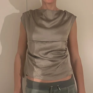 Beige ärmlös topp - Jätte fin och festlig topp från hm, helt ny aldrig använd pga storleken (köpt för 179 kr). Den är i ett siden likt material. Toppen är grå/ silvrig i färgen. Modell 1 är 178 cm och modell 2 är 155 cm! Den har en dragkedja bak så den är lätt att ta på.