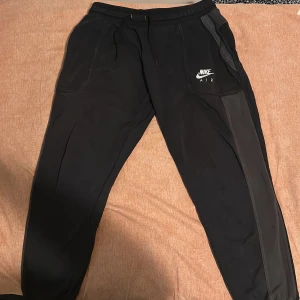 Nike mjukisbyxor - Säljer ett par svarta mjukisbyxor från Nike med elastisk midja och snörning. Byxorna har en vit Nike-logga på sidan och praktiska fickor. Perfekta för en avslappnad stil. (Byxan har ett håll)