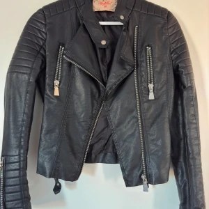 Svart skinnjacka med dragkedjor - Snygg svart skinnjacka med coola dragkedjor och quiltade detaljer på axlarna. Jackan har en klassisk bikerstil med bältesdetaljer i nederkant. Från Chiquelle i storlek 38. Den har spruckit lite baktill på ena sidan (se bild). Det är enkelt att laga för den som kan.