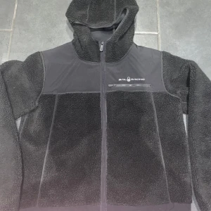 Svart fleecejacka från Sail Racing - Svart fleecejacka från Sail Racing med huva och dragkedja. Jackan är skön och är fin, är perfekt för kyligare dagar😁