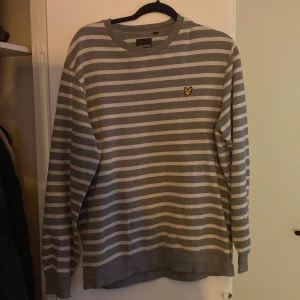 Grå och vit randig tröja från Lyle & Scott - Säljer en stilren grå och vit randig tröja från Lyle & Scott med deras ikoniska gula logga på bröstet. Tröjan har långa ärmar och en bekväm passform, perfekt för vardagsbruk.