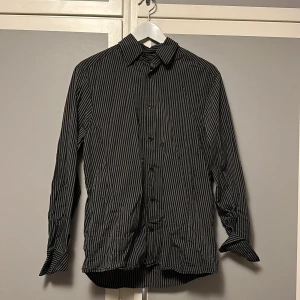 Svart och vit randig skjorta från H&M - Snygg svart och vit randig skjorta från H&M i storlek M. Skjortan har långa ärmar och knappar framtill. Perfekt för en stilren look.