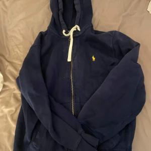 Mörkblå huvtröja från Ralph Lauren - Säljer en mörkblå huvtröja från Ralph Lauren med dragkedja och vit snörning i huvan. Tröjan har långa ärmar och en liten gul logga på bröstet. Perfekt för en avslappnad stil.