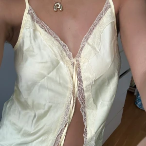 Gul linne med spetsdetaljer - Säljer ett sött gult linne med smala axelband och v-ringning. Toppen har fina spetsdetaljer längs kanterna och en knytning framtill. Perfekt för varma dagar eller som ett lager under en kofta. Är egentligen en pyjamas topp! Men funkar till allt!