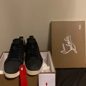 Mörkblå sneakers från Christian Louboutin - Snygga mörkblå sneakers från Christian Louboutin med den ikoniska röda sulan. Skorna har snörning och en stilren design som passar perfekt för en trendig look. Allt og medföljer