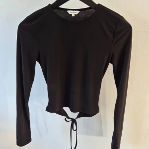 Svart långärmad topp med knyt XS - Snygg svart långärmad topp från NA-KD med knytning i midjan. Perfekt för en trendig look, passar bra till jeans eller kjol. Toppen är croppad och har en stilren design.