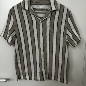 Randig kortärmad skjorta från Zara strl S - Snygg kortärmad skjorta från Zara med vertikala ränder i beige, svart och vit. Skjortan har en avslappnad passform och knappar framtill. Perfekt för en stilren look.