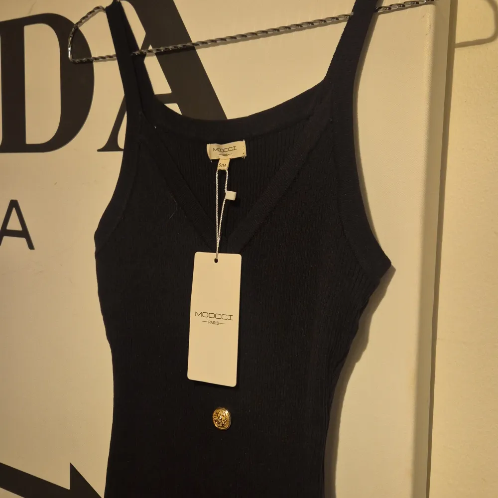 Snygg svart ribbad klänning från Moccou inköpt på raglady med guldknappar framtill. Klänningen har en tight passform och är ärmlös, perfekt för en stilren look. Den är gjord i ett mjukt material som ger en bekväm känsla.. Mekot.