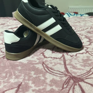 Svarta sneakers med vita detaljer - Snygga svarta sneakers med vita ränder och detaljer. Skorna har en klassisk design med snörning och en brun sula. Perfekta för en avslappnad stil.