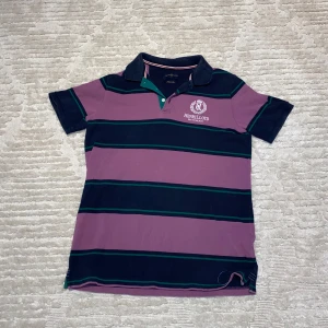 Randig pikétröja från Henri Lloyd - Säljer en snygg randig pikétröja från Henri Lloyd i lila och marinblått med gröna detaljer. Tröjan har korta ärmar och en klassisk krage med knappar. Perfekt för en avslappnad stil med en touch av brittisk segling.