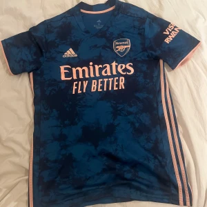 Blå Arsenal fotbollströja från Adidas - Snygg blå Arsenal fotbollströja från Adidas med korta ärmar. Tröjan har ett unikt mönster och loggor i kontrasterande färger. Perfekt för alla Arsenal-fans som vill visa sitt stöd.
