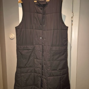 Svart quiltad väst från Pieces - Snygg svart quiltad väst från Pieces i storlek S. Västen har knappar framtill och stora fickor, perfekt för lager på lager-stil. Lång modell