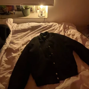 Svart bomberjacka från weekday  - Snygg svart bomberjacka med knappar och broderad text på ryggen. Perfekt för en cool och avslappnad stil.