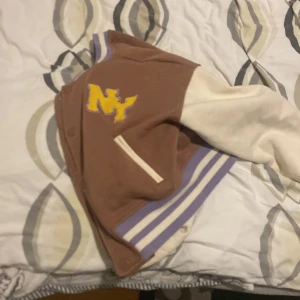 Brun hoodie med ( NY ) märket - Säljer en snygg bomberjacka i brunt och vitt med en stor gul NY-logga på bröstet. Jackan har vita ärmar och ribbade muddar i lila och vitt. Perfekt för en avslappnad stil.