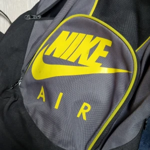 Svart och grå ryggsäck från Nike - Snygg och praktisk ryggsäck från Nike i svart och grått med gult Nike Air-tryck. Perfekt för skolan eller träningen med justerbara axelremmar och flera fack för förvaring.