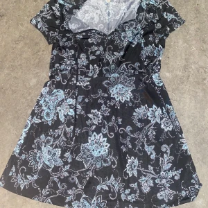 Svart blommig blus - Charmig svart blus med ett vackert blommigt mönster i ljusblått och vitt. Klänningen har korta ärmar och en omlott-design som ger en elegant touch. Perfekt för både vardag och speciella tillfällen.