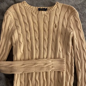 Beige kabelstickad tröja från Ralph Lauren - Snygg beige kabelstickad tröja från Ralph Lauren. Den har en klassisk rund halsringning och långa ärmar. Perfekt för kyligare dagar och ger en stilren look.