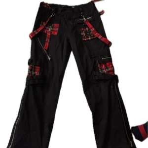 Svarta cargopants med röda rutiga detaljer - Unika svarta cargopants med coola röda rutiga detaljer och dragkedjor. Byxorna har flera fickor och justerbara remmar för en edgy look. Perfekta för dig som vill sticka ut!