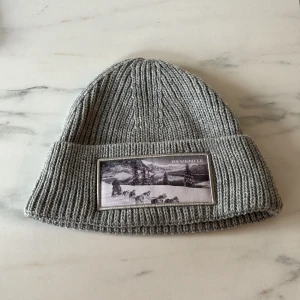 Grå ribbad mössa med broderad patch - Snygg grå ribbad mössa med en broderad patch framtill. Perfekt för kyliga dagar och ger en stilren look. Mössan är mjuk och bekväm att bära.