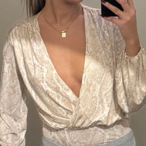 Beige omlottbody - Snygg beige omlottbody med ormmönster från NLY One. Toppen har långa ärmar och en djup v-ringning. Materialet är mjukt och bekvämt. Använd 1 gång.