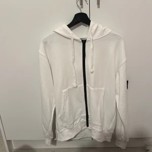 Vit hoodie från Stone Island - Säljer en stilren vit hoodie från Stone Island med dragkedja och justerbar huva. Tröjan har en ikonisk logga på ärmen och praktiska fickor framtill. Perfekt för en avslappnad look.