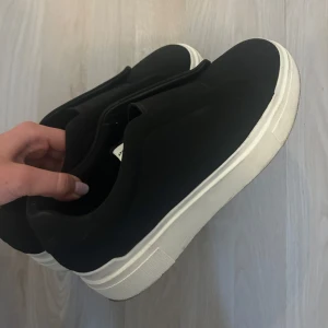 Svarta sneakers - Säljer ett par stilrena svarta sneakers med vit sula. Skorna har en enkel design utan snörning, vilket gör dem lätta att ta på och av. Väldigt snygga att styla outfits med men tyvärr så är de nån aning förstora för mig. Använda Max 3 ggr och kommer att tvättas av innan de skickas iväg! Dessa säljs dessutom inte längre!