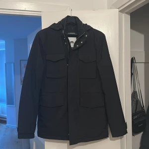 Svart jacka från J.Lindeberg - Snygg svart jacka från J.Lindeberg med huva (går att dölja) och flera praktiska fickor. Jackan har dragkedja och knappar framtill för extra skydd. Perfekt för höst och vår. Storlek S och passar mellan 1,75 och 1,82 lång skulle jag säga