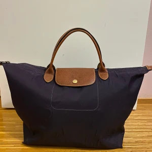 Longchamp väska lila  - Snygg lila axelväska från Longchamp med bruna läderdetaljer och dragkedja. Perfekt för både vardag och resa med sin rymliga design och stilrena look. Den har blivit lite skrynklig av vatten som syns på bild 4 och den har väldigt små hål i hörnen men inget som stör funktionen💕💜