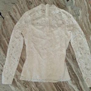 Vit spetsblus från Vila - Elegant vit spetsblus från Vila med långärmad design och hög hals. Blusen har ett vackert spetsmönster och en dragkedja i ryggen för enkel påklädning. Perfekt för en stilren look. Orginalpris: 400 kronor