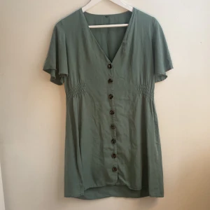 Grön klänning med knappar - Snygg grön klänning med v-ringning och korta ärmar. Den har dekorativa knappar framtill och smockdetaljer vid midjan för en smickrande passform. Perfekt för en avslappnad stil. Väldigt svalt material.