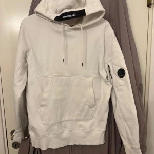 Vit hoodie från C.P. Company - Snygg vit hoodie från C.P. Company med en klassisk känguruficka och justerbar huva. Den har en unik detalj med en rund emblem på ärmen. Perfekt för en stilren och avslappnad look. Inga fläck eller hål! Storlek L men passar även M