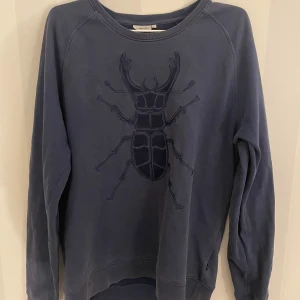 Blå tröja från J.Lindeberg - Säljer en blå tröja från J.Lindeberg med ett stort skalbagge-motiv på framsidan. Tröjan har långa ärmar och en rund halsringning. Perfekt för en avslappnad stil. Tveka inte att hojta till vid eventuella frågor!