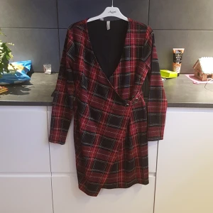 Rutig omlottklänning från Bodyflirt - Snygg rutig omlottklänning från Bodyflirt i rött och svart med inslag av vitt. Klänningen har en v-ringad design och knappar framtill. Perfekt för en stilfull look!