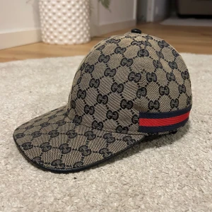 Gucci keps - Snygg keps från Gucci med det ikoniska GG-mönstret. Kepsen har en röd och blå randdetalj på sidan och justerbart spänne baktill. Perfekt för att ge din outfit en lyxig touch. Kepsen är i mycket bra skick och är storlek S men kan även passa dig som har M. Skriv för intresse! 
