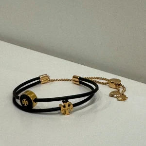 TORY BURCH armband - Kira Slider i färgen guld och svart - Kira Bracelet är inspirerad av retro vänskapsstilar. Man kan justera skjutreglaget för att få en perfekt passform.  - Läder, 18k guldpläterad mässing, slipad emalj - Justerbart skjutreglage - Nickelfri