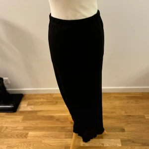 Svart långkjol från NA-KD - Säljer en elegant svart långkjol från NA-KD. Kjolen har en stilren design med en elastisk midja för extra komfort. Perfekt för både vardag och festliga tillfällen. Materialet känns mjukt och följsamt, vilket gör den skön att bära hela dagen.