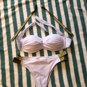 Vit bikini med gulddetaljer - Snygg vit bikini med guldfärgade banddetaljer. Överdelen har en bandeau-stil med knytband och avtagbara axelband. Underdelen har matchande gulddetaljer på sidorna för en extra touch av glamour.