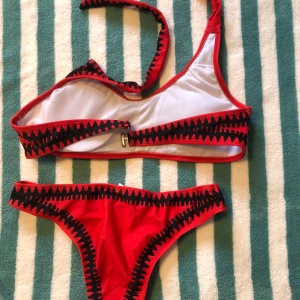 Röd och svart bikini med zigzag-mönster - Snygg röd bikini med svart zigzag-mönster. Toppen har en unik design med knytband och en liten rosett framtill. Perfekt för en dag på stranden eller vid poolen!